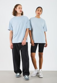 Lys blå oversized t-skjorter båret av en mann og en kvinne, sammen med svarte sykkelshorts og joggesko. Bakgrunnen er helt hvit.