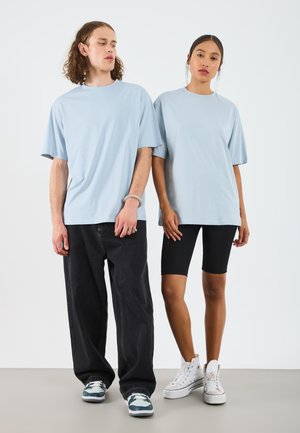 Hellblaue, übergroße T-Shirts, die von einem Mann und einer Frau getragen werden, kombiniert mit schwarzen Radshorts und Turnschuhen. Der Hintergrund ist schlicht weiß.