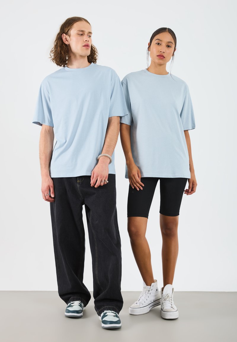Lys blå oversized t-skjorter båret av en mann og en kvinne, sammen med svarte sykkelshorts og joggesko. Bakgrunnen er helt hvit.