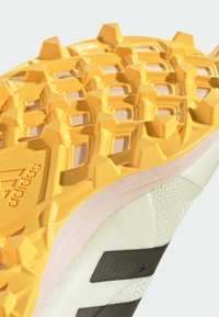 Sula på en sko med en texturerad gul gummiyttersula med vinklade spår, vit mellansula och en mönstrad övre del i ljusgrå.