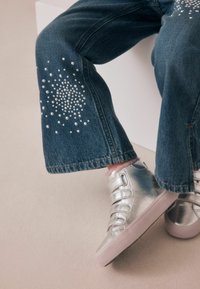 Jeans med utsvängda ben och silverfärgade nitar, parat med metalliska silverfärgade sneakers med elastiska remmar och en strukturerad sula.