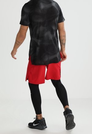 Personne debout de dos portant un t-shirt camouflage noir, un short rouge par-dessus un legging noir et des baskets Nike noires sur fond blanc.