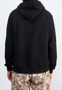 Osoba viděná zezadu, oblečená v jednoduché černé mikině a béžových kalhotách s mnohobarevným leopardím potiskem.