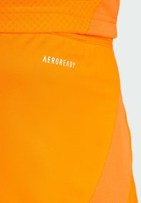 Oranssi urheilushortsit, joissa on sileä pinta ja verkkokangasvimma. "AEROREADY"-logo on painettu valkoisena kankaaseen.