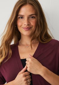Bordeaux gebreide cardigan met een V-hals en ribbelstructuur, gedragen over een zwarte top. Model heeft lang haar en eenvoudige zilveren sieraden.
