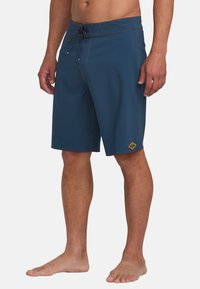 Pantaloni da bagno blu con coulisse, cuciture gialle a contrasto e un'etichetta con logo. Il design è lungo fino al ginocchio ed è realizzato in tessuto liscio.