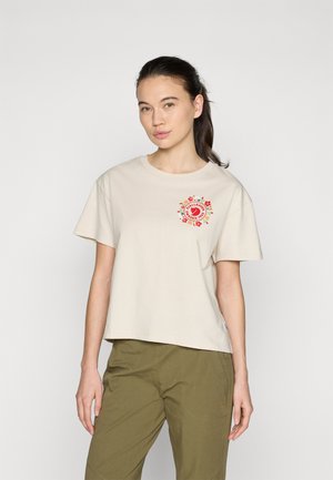 Beige t-paita lyhyillä hihoilla, jossa on punainen kukkainen sydänkuvio rinnassa. Yhdistetty oliivinvihreisiin housuihin.