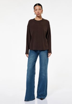 Vrouw met gouden oorringen, een donkerbruin shirt met lange mouwen en wijde blauwe jeans, staand tegen een witte achtergrond.