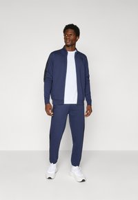 Fato de treino desportivo da Marinha com casaco com fecho éclair e calças com cintura elástica; realces pretos contrastantes nas mangas do casaco e nas laterais das calças; t-shirt branca por baixo.