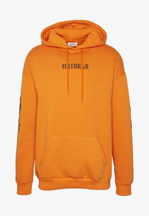 Persoon staat met gekruiste armen, draagt een zwarte puffer-vest over een oranje hoodie met rozenprint, donkere jeans en zwarte veterlaarzen.