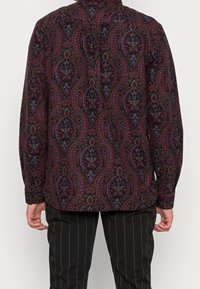 Long-sleeve shirt in dark fabric with a colorful paisley pattern, featuring a collar and buttoned cuffs, paired with pinstriped pants.