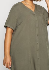Abito camicia verde oliva con bottoni realizzato in un tessuto morbido, dotato di scollo a V, maniche corte e tasca frontale.