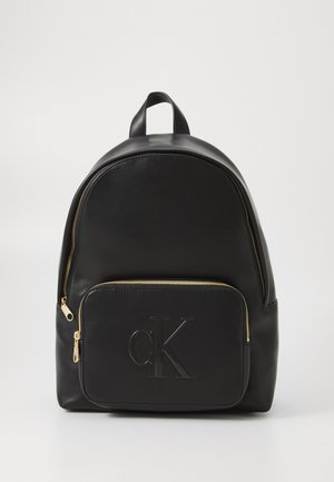 Mochila de cuero negra con forma redondeada, cremallera dorada y un bolsillo frontal con un logotipo "ck" en relieve. Textura suave en todo.
