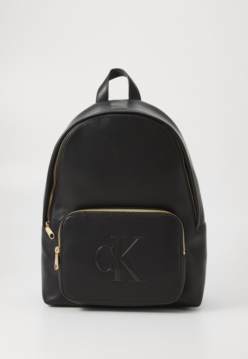 Rucsac din piele neagră cu o formă rotunjită, fermoar auriu și un buzunar frontal cu un logo "ck" aplicat. Textură netedă pe toată suprafața.