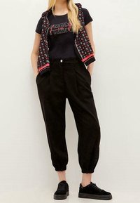 Zwarte trainingsbroek met cuffed enkels en zijzakken, gecombineerd met een zwart T-shirt met rode letters en een gepatternde vest. Zwarte sneakers.