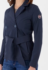 Giacca doppiopetto blu navy con vita cintata, realizzata in tessuto liscio. Presenta due bottoni blu e una toppa con logo sulla spalla.