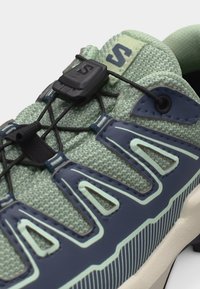 Κοντινή λήψη ενός πράσινου και ναυτικού αθλητικού παπουτσιού με υφασμένο επάνω μέρος και σύστημα γρήγορου δέσιμου με μαύρο κούμπωμα και λογότυπο.