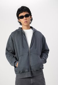Gray zip-up hoodie made of soft fabric, featuring a drawstring hood, front pockets, and ribbed cuffs. Worn over a white T-shirt.