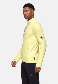 Lichtgele zip-up jas gemaakt van zachte stof, met een hoge kraag, twee zijzakken en een cirkelvormig logo op de linkermouw.