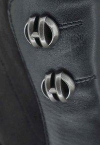 Primo piano di un indumento in pelle nera con due bottoni metallici ovali dal design stratificato e una cucitura sottile lungo la cucitura.