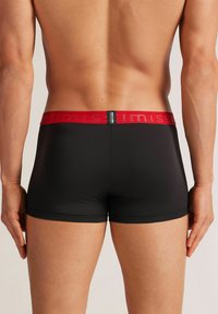 Černé boxerky s červeným elastickým pasem, na kterém je uvedeno jméno značky. Materiál vypadá hladce a těsně přiléhá.