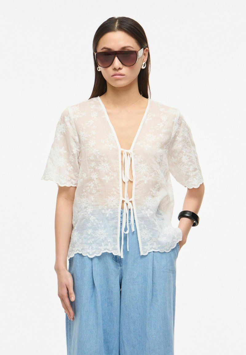 Femme portant de grandes lunettes de soleil, un chemisier blanc transparent à fleurs noué devant, un jean large bleu clair et un bracelet noir, debout sur fond blanc.