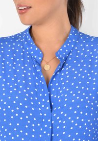 Blå blus med polkagubbar och mandarinkrage, som har vita prickar och ett guldhalsband med en cirkulär berlock. Slät textur.