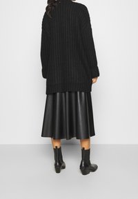 Cárdigan abierto de punto negro con textura acanalada, combinado con una falda acampanada de cuero sintético negro y botas de tobillo. Diseño simple y elegante.