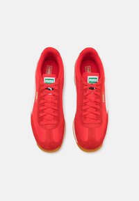 Paire de baskets rouges Puma avec des bandes blanches sur le côté, semelles en gomme, étiquettes du logo Puma vertes et blanches sur la languette, et lacets sur le devant, vues de dessus.