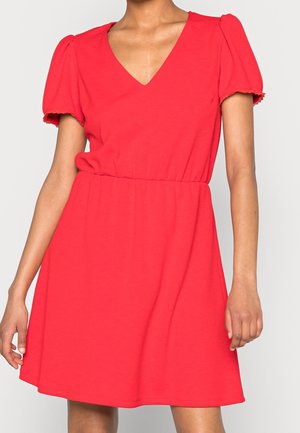 Kvinde iført en rød kortærmet kjole med V-hals og elastik i taljen, stående foran en ensfarvet lys baggrund.