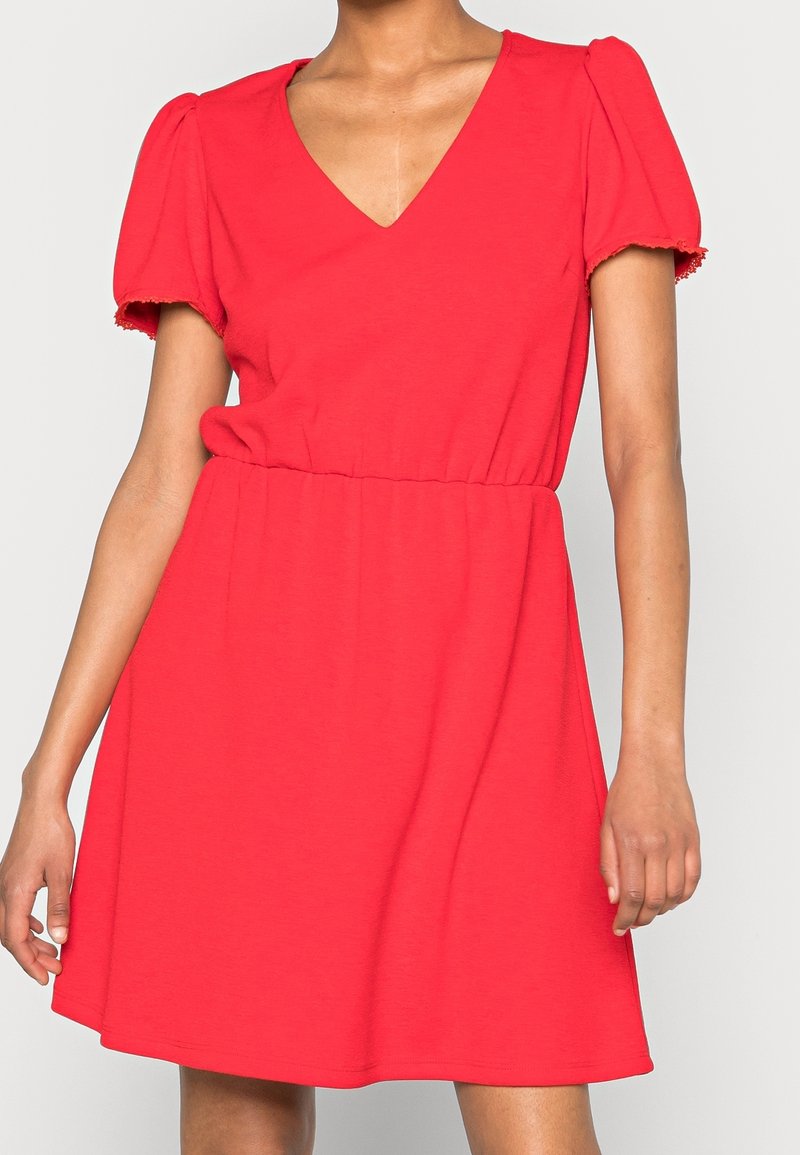 Femme portant une robe rouge à manches courtes avec un décolleté en V et une taille élastique, debout devant un fond clair uni.