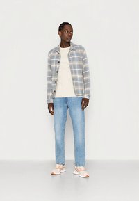 Rutete skjorte med knapper i lys grå og blå, båret over en kremfarget t-skjorte, kombinert med lyseblå jeans og beige joggesko.