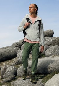 Lichtgrijze ritssluiting jas, roze t-shirt, groene cargobroek en bruine wandelschoenen. Staande op rotsen, met een rugzak in de hand, in de buitenlucht.