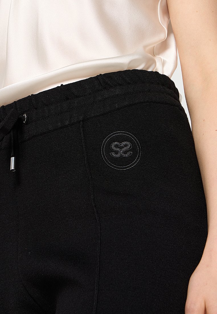 Schwarze Jogginghose mit elastischem Bund, ausgestattet mit einem Kordelzug und einem runden Logoakzent in Grau an der Hüfte. Glatte Stofftextur.
