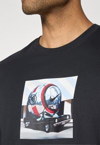 Sort bomulds t-shirt med et farverigt grafisk design af en fodbold på en vintage bil. Designet inkluderer røde og sølvfarvede accenter.