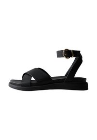 Sandal de cuero negro con correas cruzadas, hebilla en el tobillo, suela plana y acabado texturizado. Diseño minimalista con una silueta elegante.