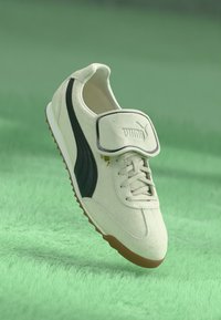 Hvide ruskinds sneakers med en grøn stribe, snørelukning, justerbar rem og brun gummisål. Puma-logoet vises på remmen.