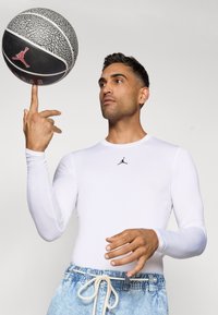 Hvid langærmet skjorte med et sort Jordan-logo, parret med lyseblå denimshorts, mens en basketball snurrer på hans fingertip.