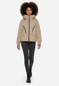 Giacca puffa beige con cappuccio, dotata di chiusura con zip, due tasche laterali e design trapuntato. Indossata con pantaloni skinny neri e stivali.