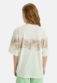 Cream-colored oversized t-shirt made of soft fabric, featuring a floral design in gold on the back. Short sleeves and a crew neckline.