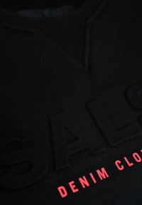 Sudadera negra con letras en relieve "OFERTA" y "ROPA DE DENIM" en rosa. Diseño con escote en V y tejido texturizado. Vista cercana.