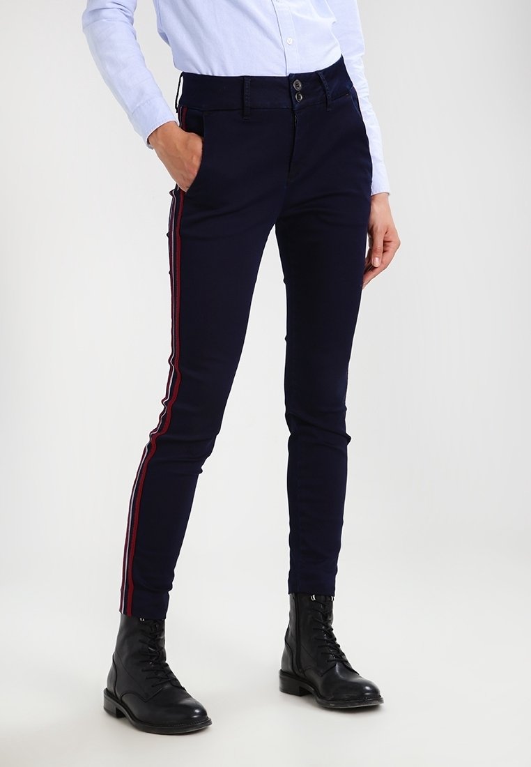 Femme portant un pantalon bleu marine ajusté avec des bandes latérales rouges et blanches, une chemise à manches longues bleu clair boutonnée, des bottines noires à lacets, une main dans la poche.