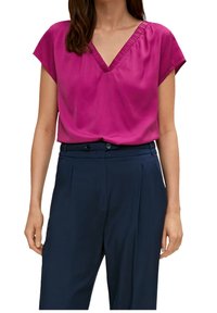 Blouse fuchsia à manches courtes avec un col en V et des plis décoratifs au niveau de l'encolure, associée à un pantalon taille haute bleu marine.