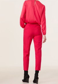 Vrouw in felroze op maat gemaakte broek en bijpassende blouse met lange mouwen, gecombineerd met zwarte haklaarzen, staande voor een effen achtergrond.