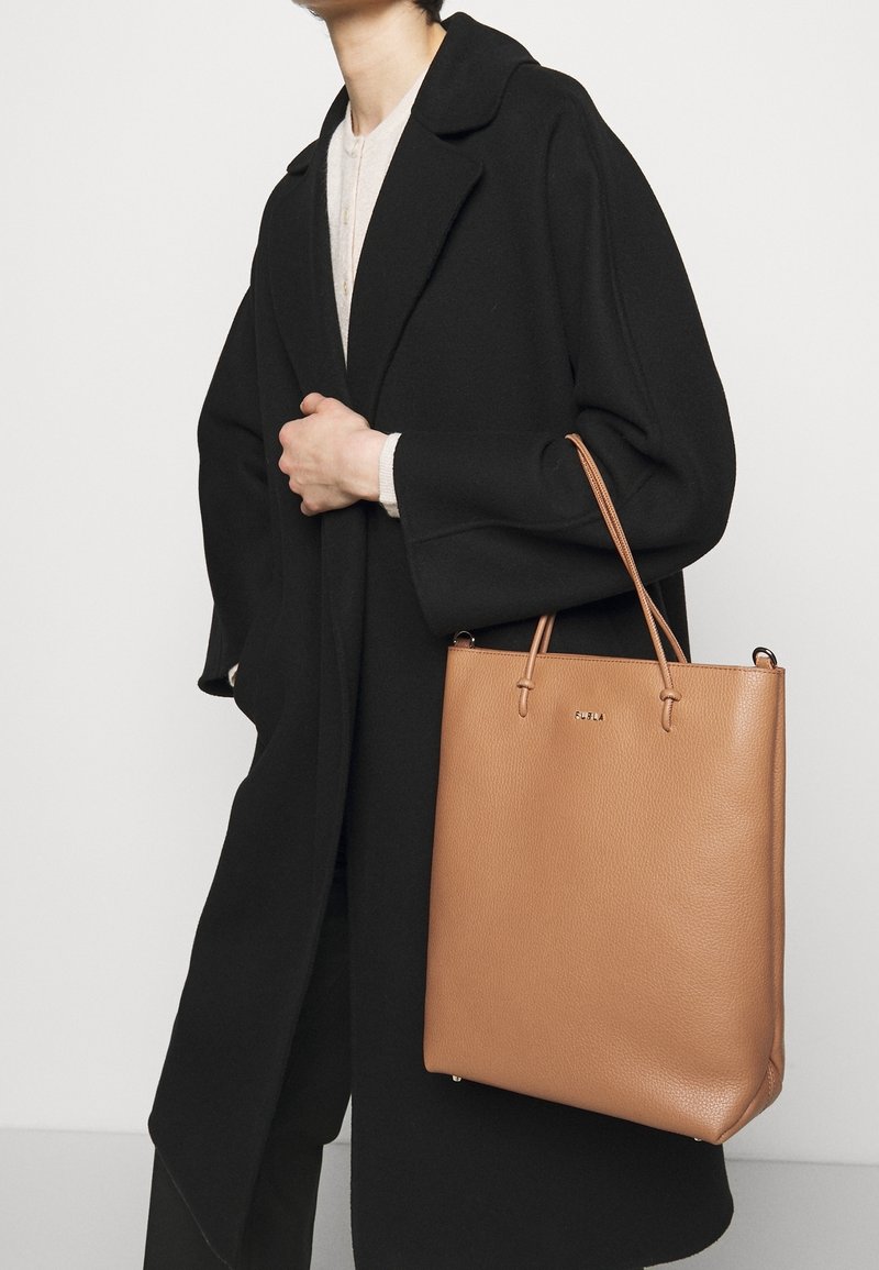 Borsa a spalla in pelle tan con una superficie testurizzata, doppi manici e un branding sottile. Indossata con un cappotto oversize nero e un top chiaro.