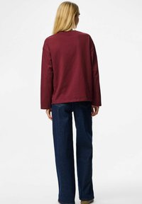 Bordowa koszula z długim rękawem i okrągłym dekoltem, w połączeniu z luźnymi, ciemnoniebieskimi dżinsami. Koszula ma prosty brzeg i swobodny krój.