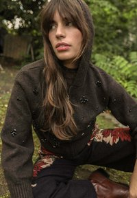 Un sweat à capuche en tricot marron avec une fermeture éclair sur le devant et des embellissements en perles florales, associé à un pantalon sombre à motifs, le tout sur fond naturel.