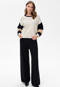 Crèmekleurige trui met zwart- en beige patronen op de mouwen, gecombineerd met zwart wijde pijpen broek met hoge taille en zwarte puntige schoenen.