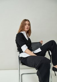 Čierny sveter s výstrihom do V, ktorý je vrstvený nad bielou košeľou s golierom, doplnený voľnými čiernymi nohavicami. Model sedí na chromovej stoličke pred neutrálnym pozadím.