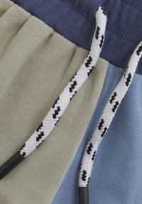 Färgblockerat tyg i beige, blått och navy; har texturerade dragsnören med svarta mönster och svarta plastspetsar. Slät stickad textur.